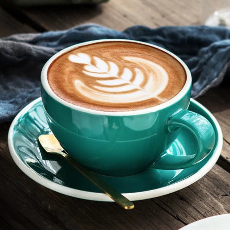 A teal ceramic cup filled with latte art sits on a matching saucer on a rustic wooden table, with a small green radio, blue cloth, flowers, and a dessert in the background.