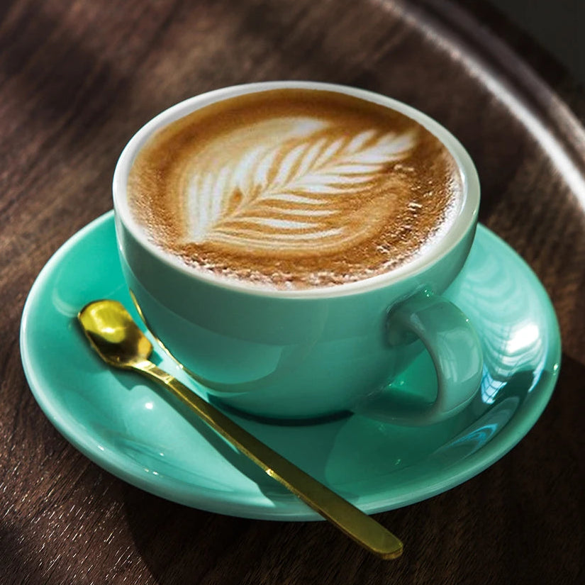 A turquoise ceramic coffee cup filled with latte art sits on a matching saucer with a gold spoon, placed on a dark wooden tray in warm sunlight.