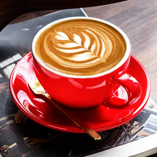 A red ceramic coffee cup and matching saucer holding a latte with leaf latte art, styled on top of an open magazine with a wooden guitar body in the background.