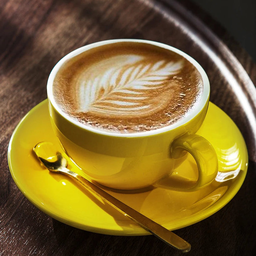 A bright yellow ceramic coffee cup filled with latte macchiato sits on a matching yellow saucer with a gold spoon, placed on a dark wooden tray.