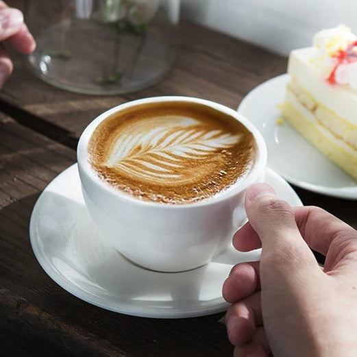 A white ceramic coffee cup filled with latte art sits on a wooden table, held by a hand. A slice of layered cake is on a plate nearby, with a glass vase of small white flowers in the background.